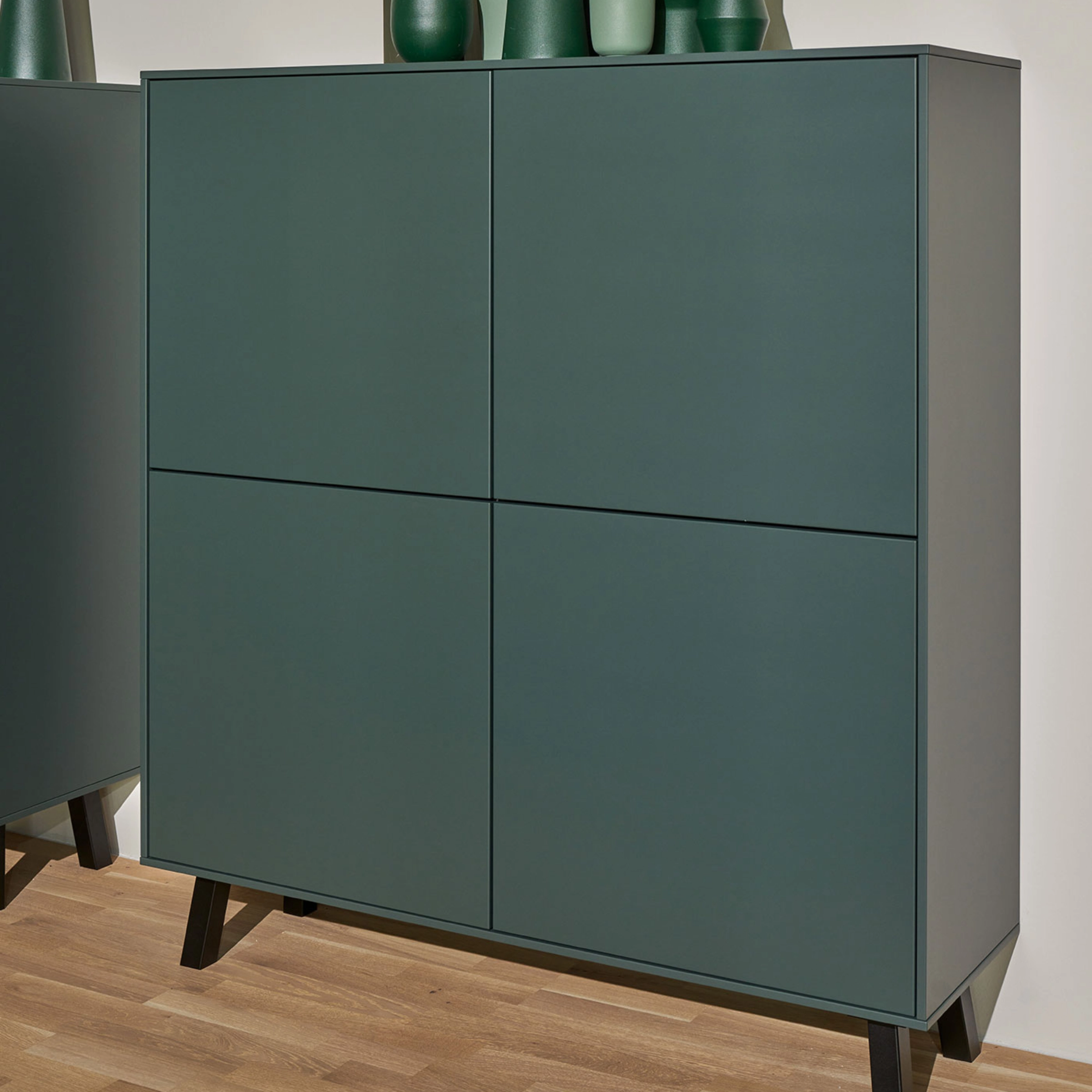 Une armoire verte avec des vases décoratifs dessus se tient sur un sol en bois. Nolte Living.