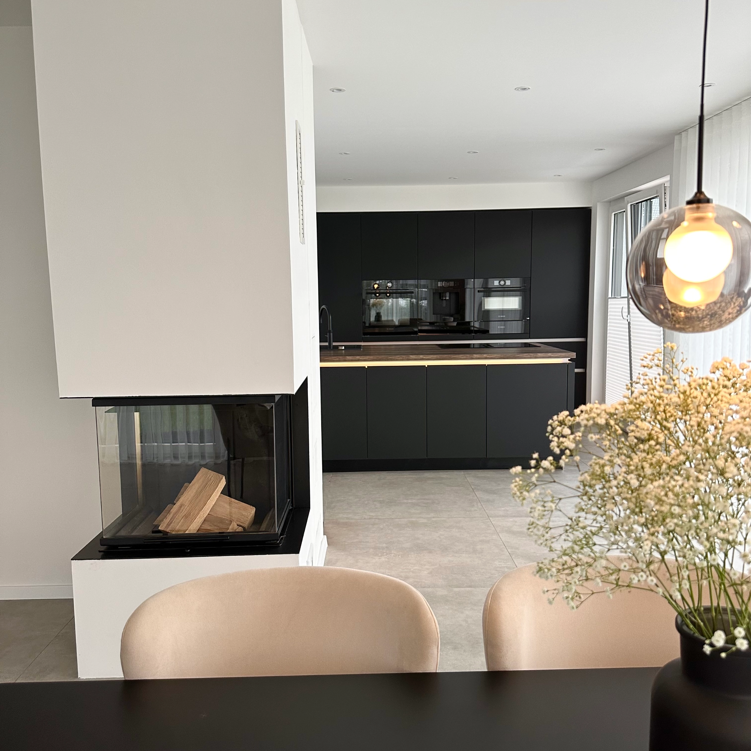 A modern kitchen with black decor and a fireplace, dining table in the foreground. Nolte Kitchens.