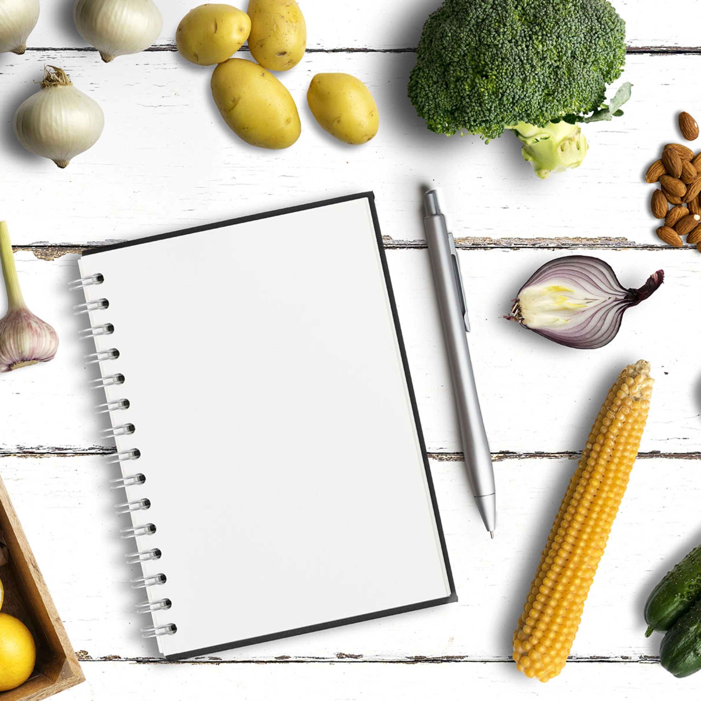 Un carnet avec un stylo entouré de légumes sur une table en bois blanc.