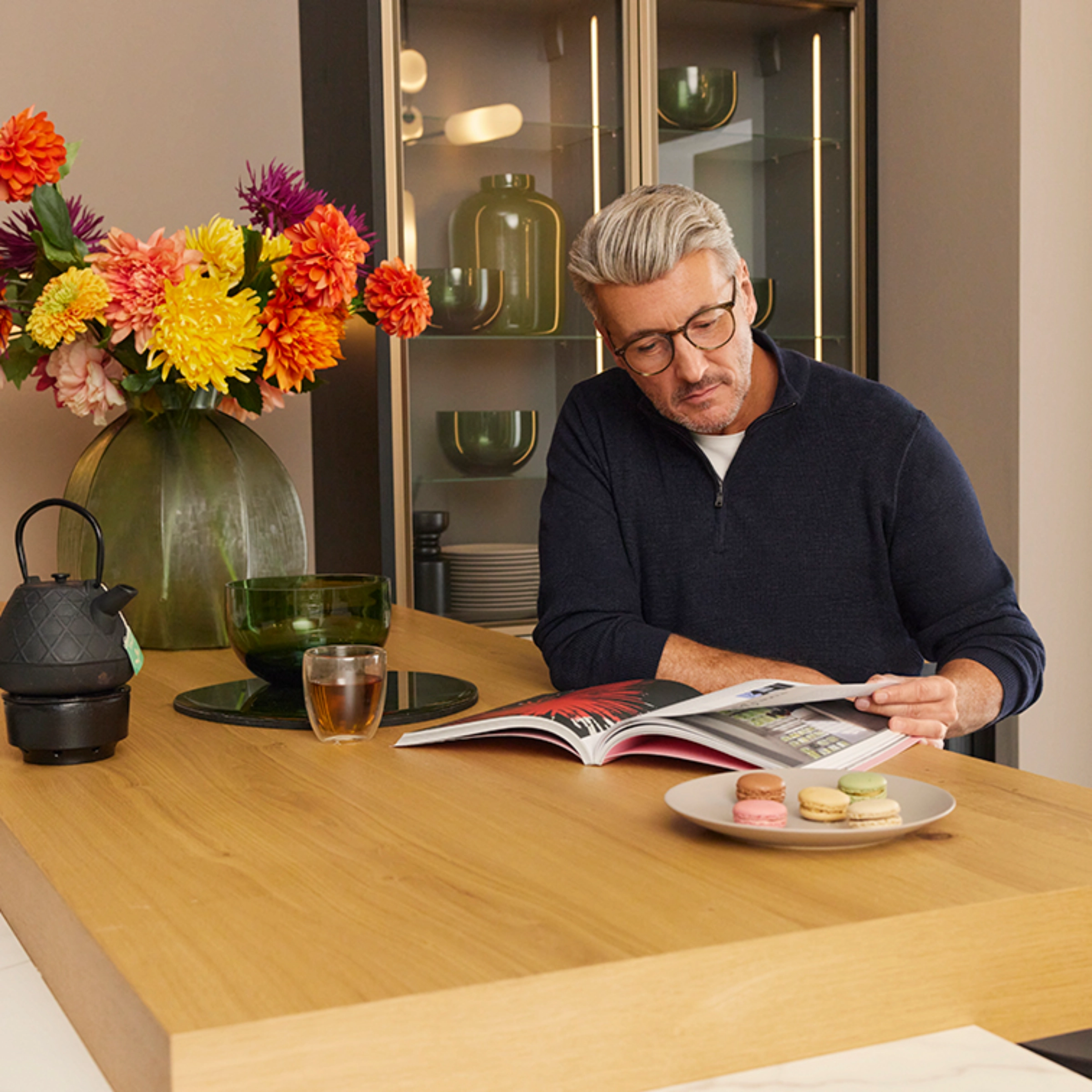 A man sits at a kitchen counter with flowers and macarons, reading a magazine. Nolte Kitchens.