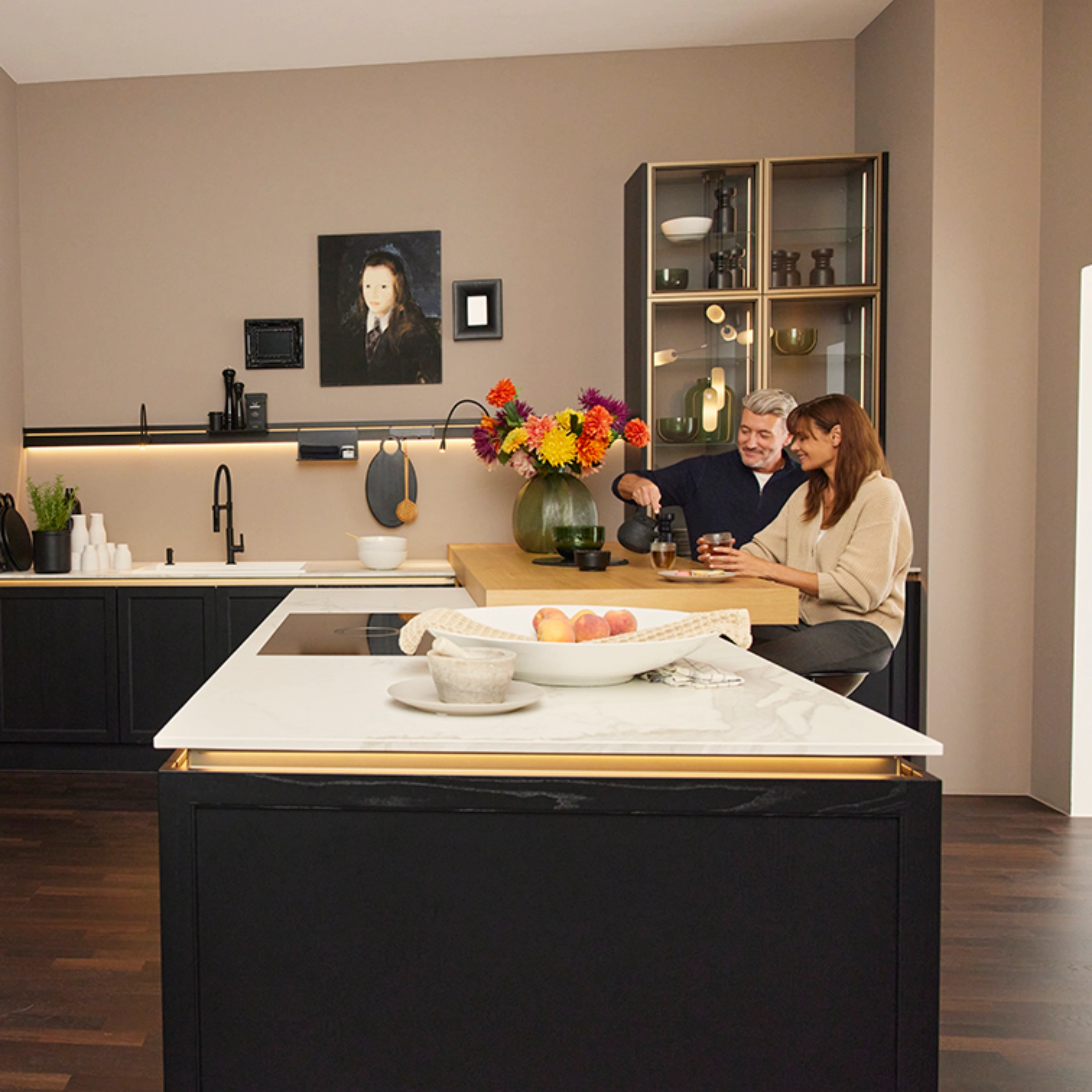 A couple sits in a modern kitchen with dark cabinets and flowers, Nolte Kitchen.