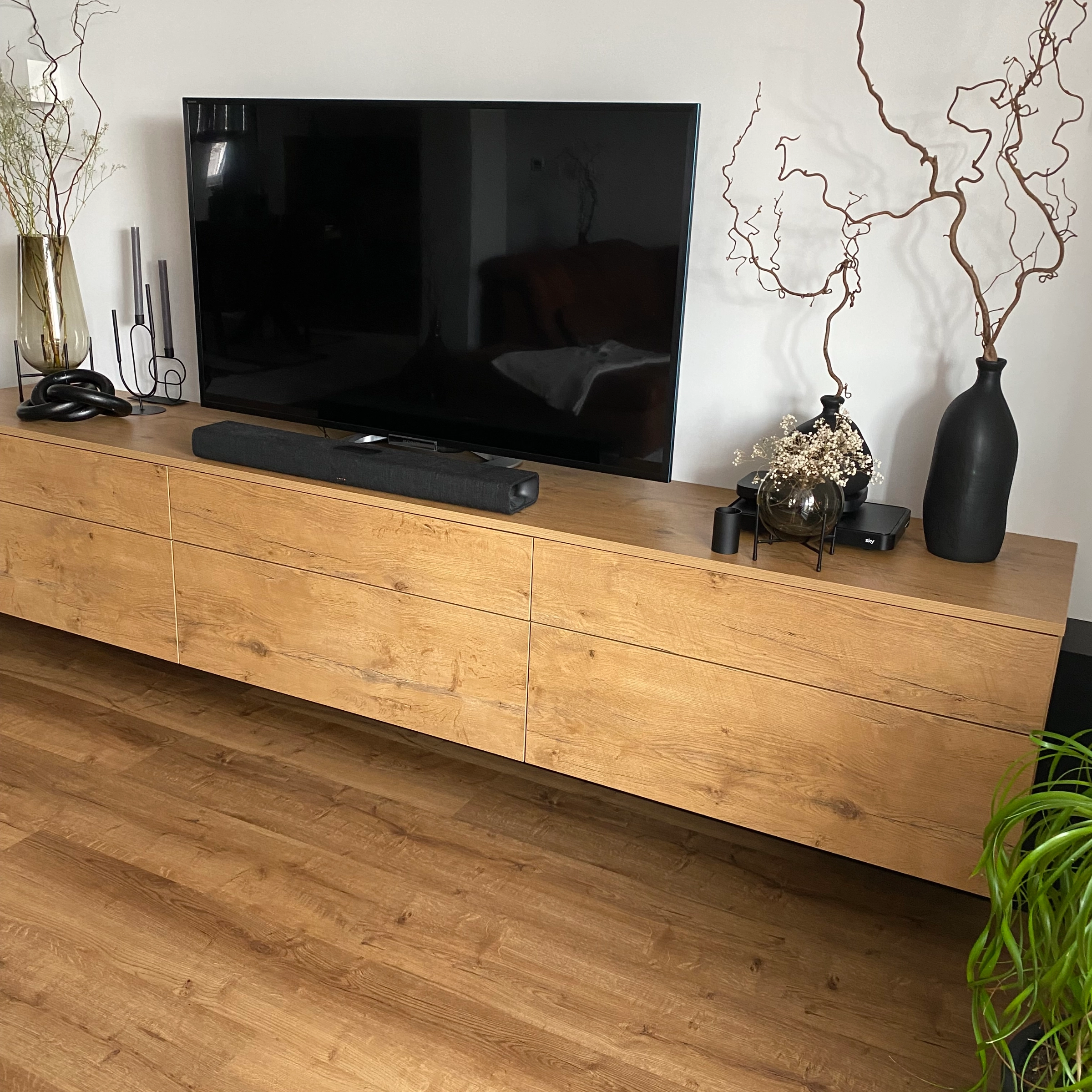 Un salon moderne avec un buffet en bois, décoré de vases et plantes, et une grande télé dessus. Nolte Living.