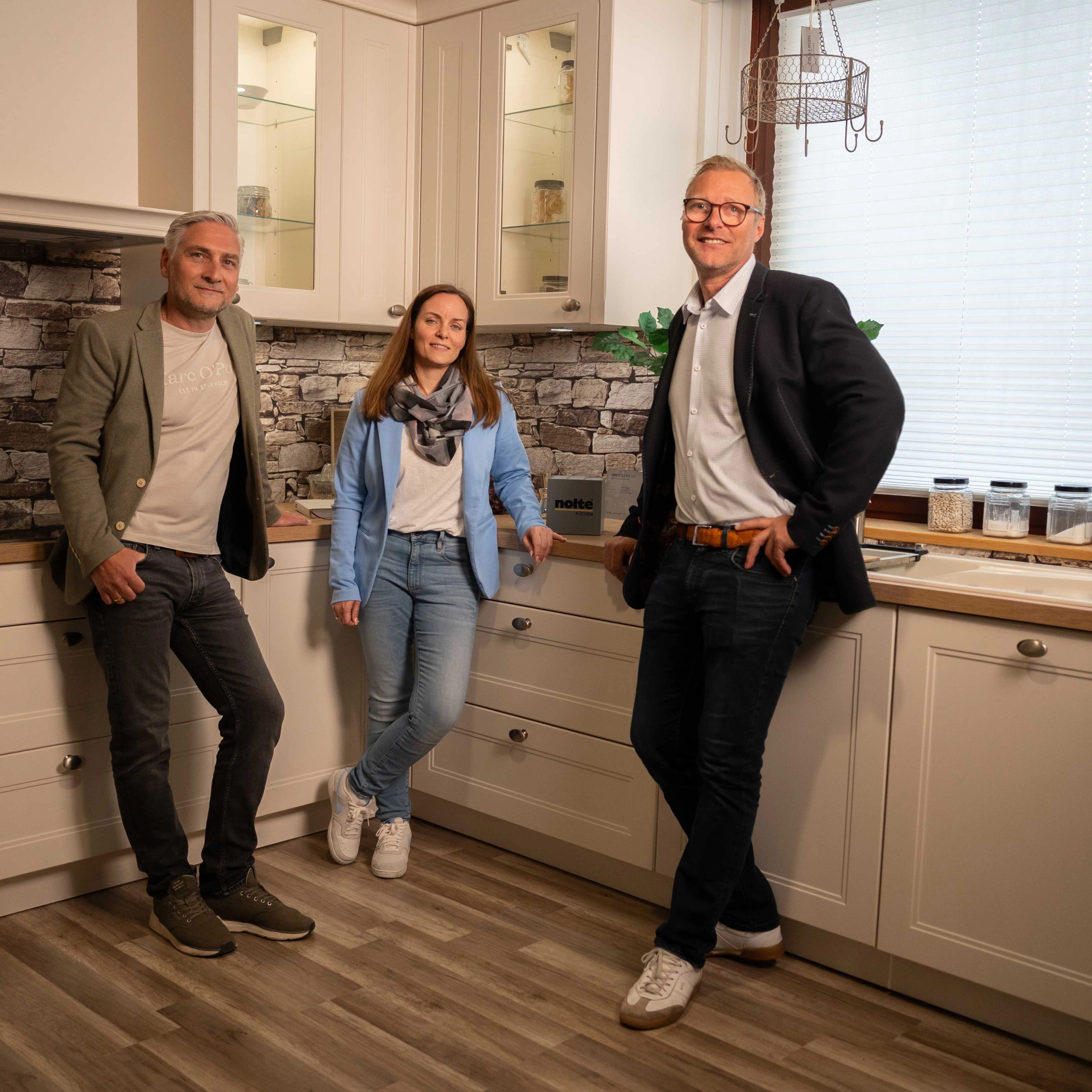 Tres personas en una cocina moderna con pared de piedra y suelo de madera. Nolte Cocina.