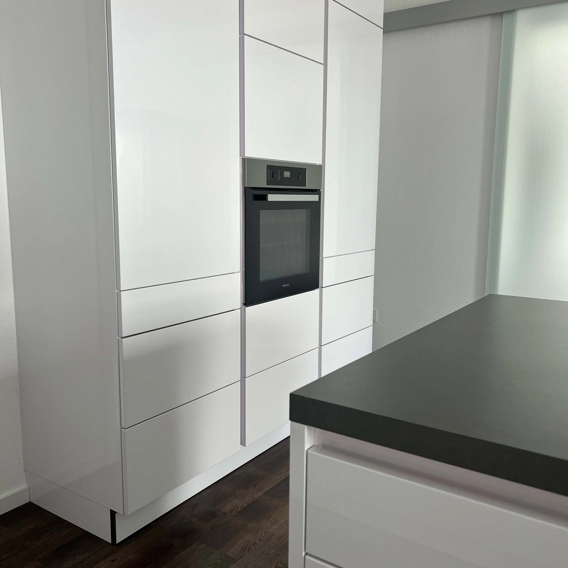 Cuisine moderne blanche avec four encastré et sol en bois foncé, conçue par Nolte Kitchen.