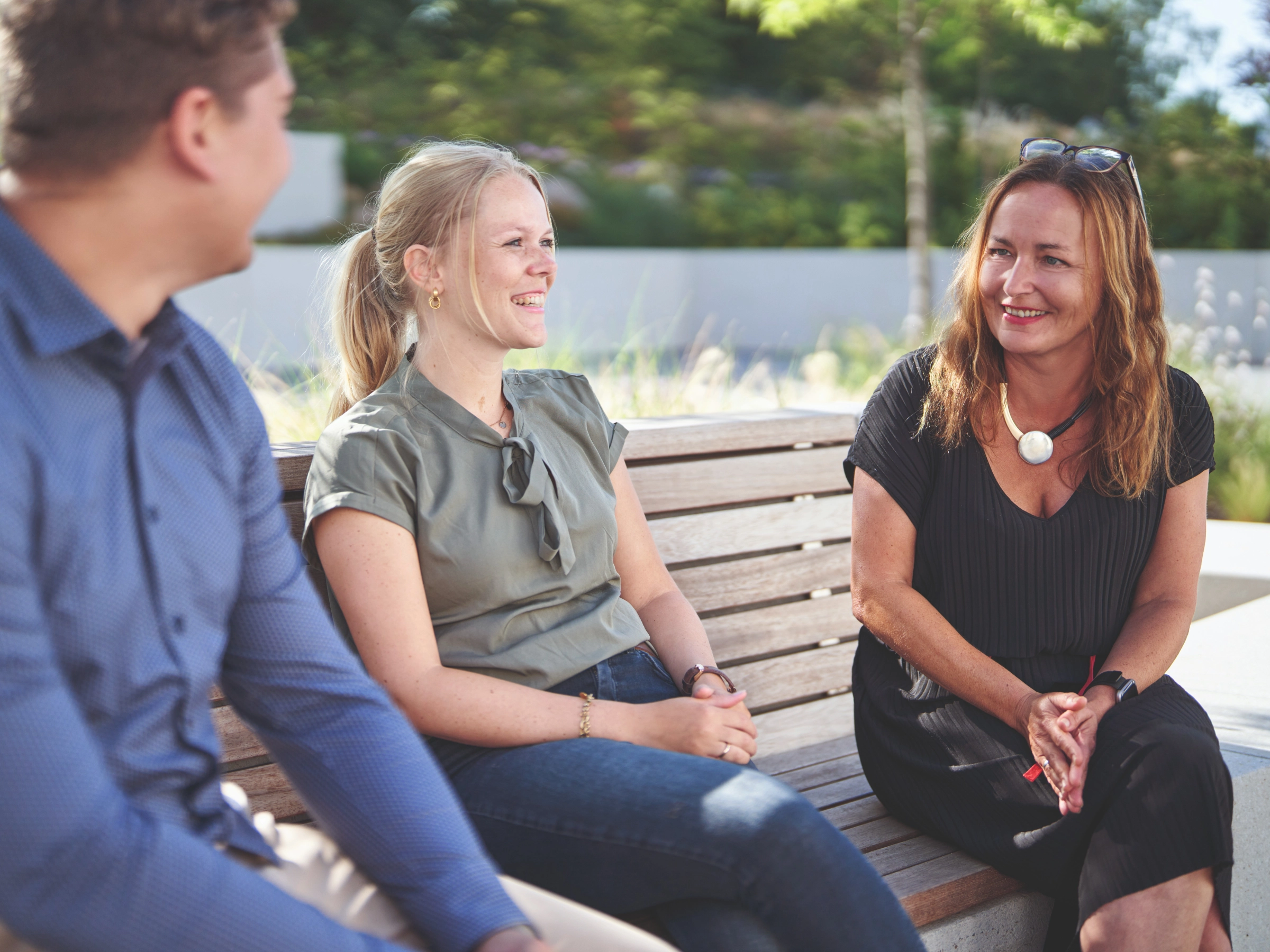 Trois personnes assises sur un banc à l'extérieur discutent par temps ensoleillé.