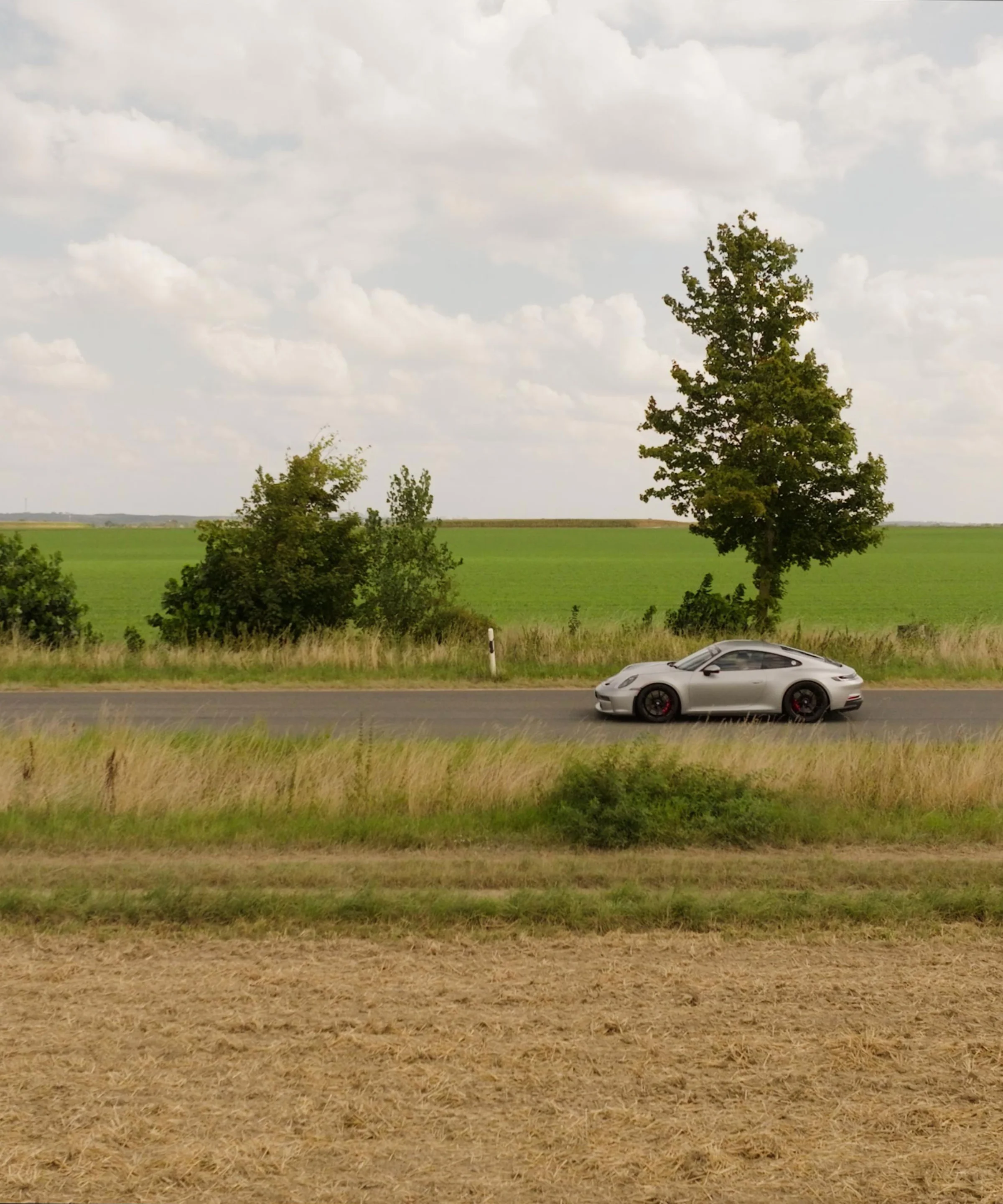 Une voiture roule sur une route de campagne bordée de champs verts sous un ciel nuageux.