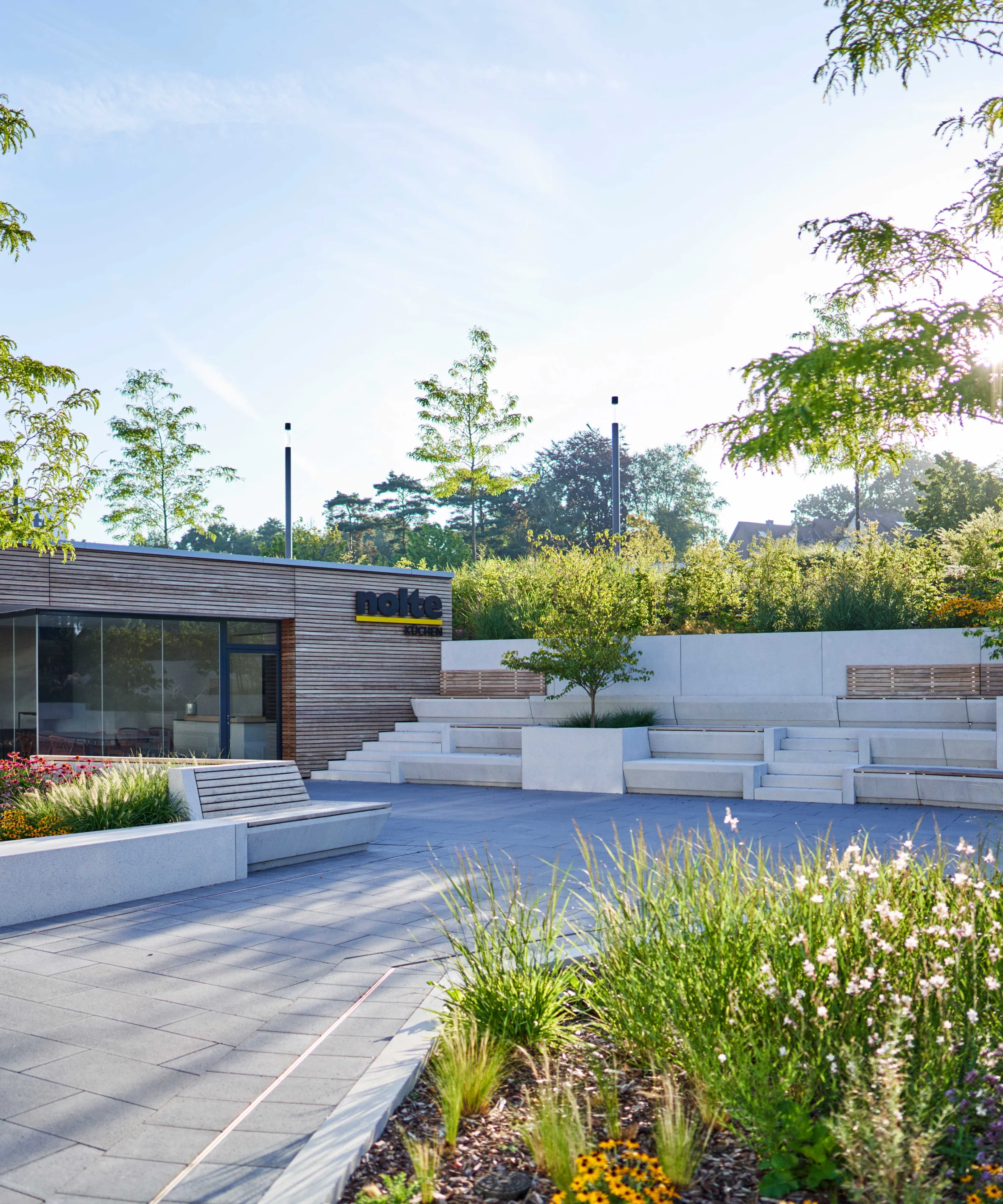 A modern outdoor area with seating and plants, with a building featuring Nolte Kitchens in the background.