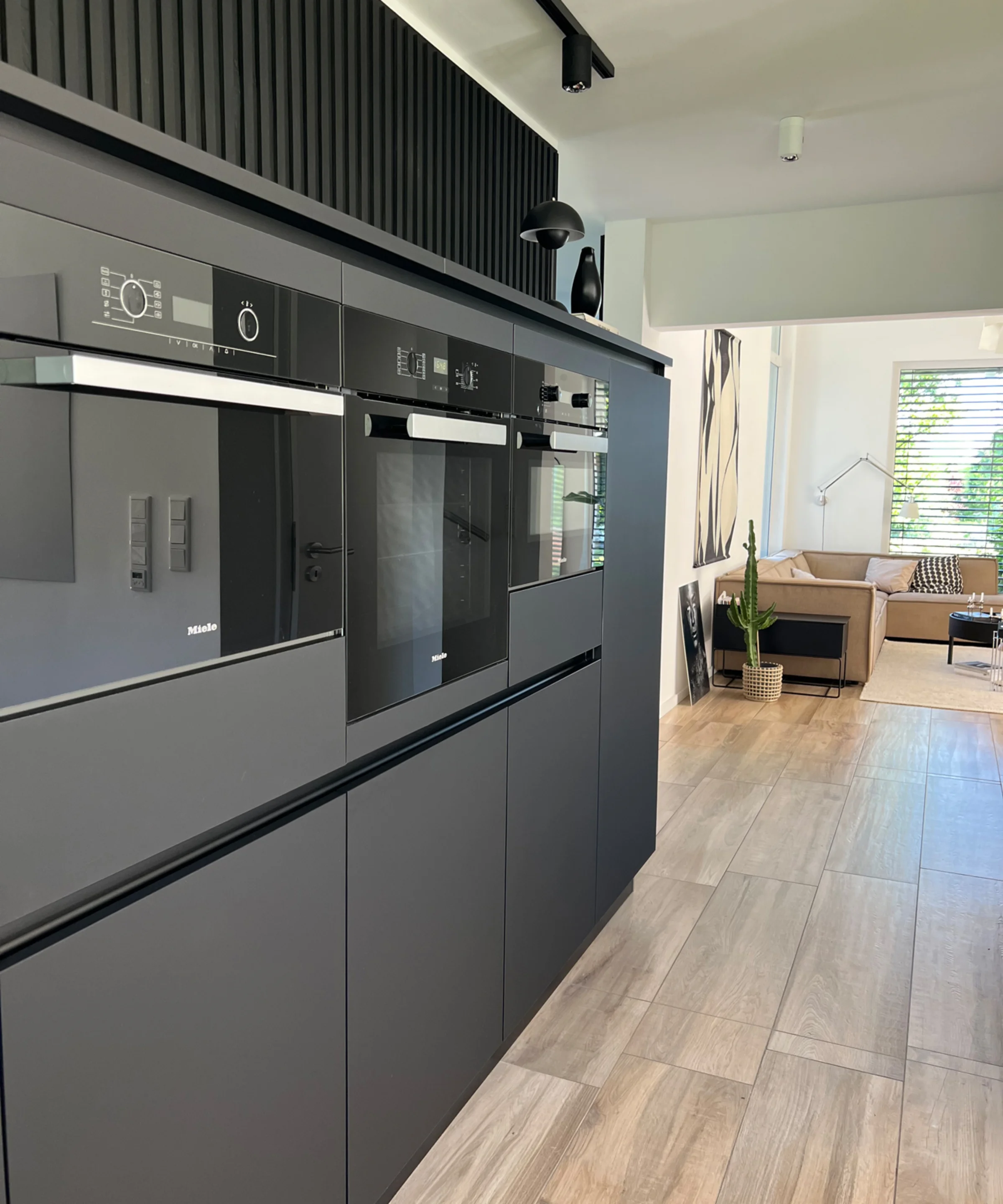 Eine moderne Küche mit schwarzen Einbaugeräten und Holzfußboden, im Hintergrund ein Wohnzimmer. Nolte Küchen.
