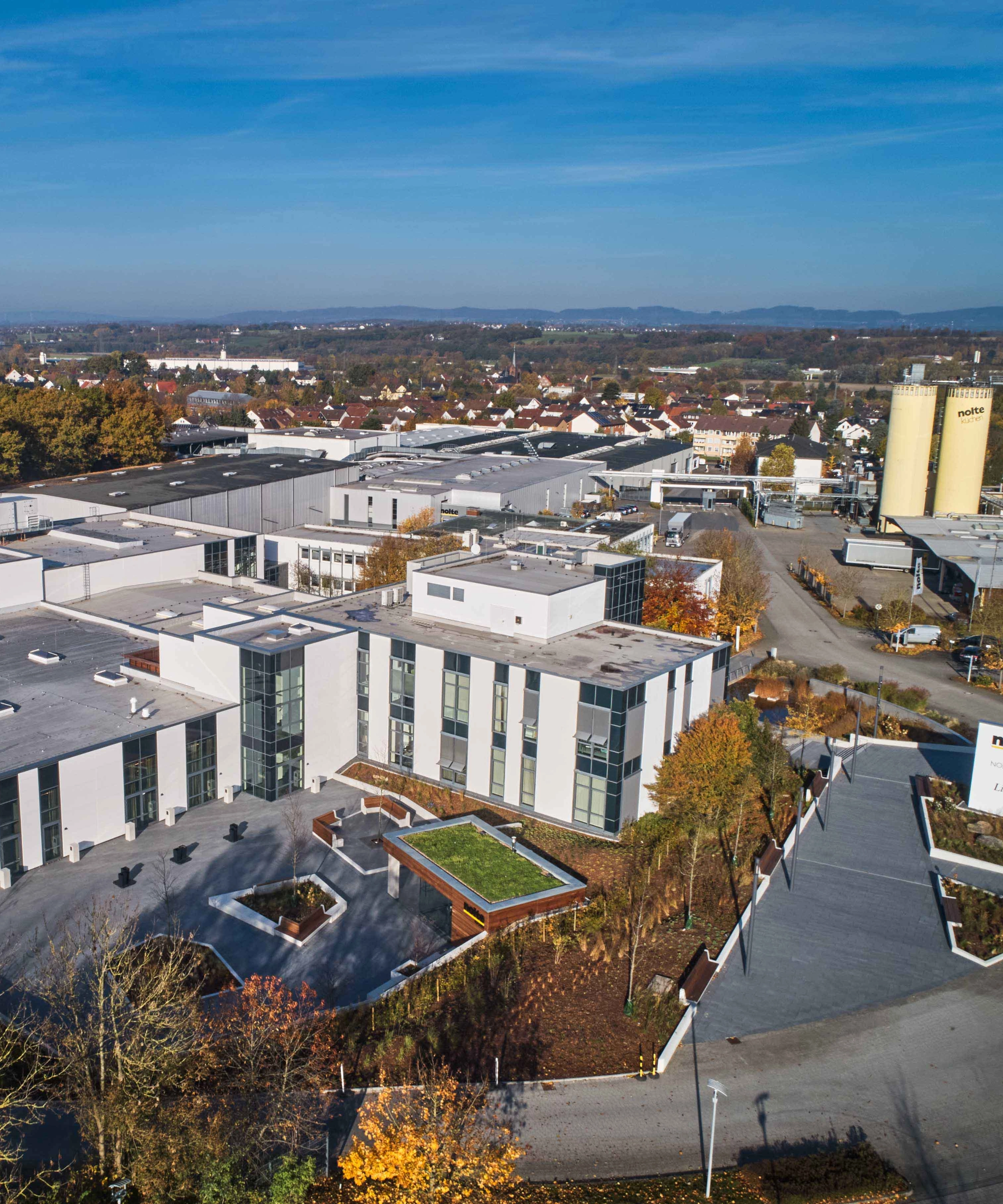 Ein modernes Gebäude mit großen Glasfronten, umgeben von einer herbstlichen Landschaft, ist zu sehen. Nolte Küchen.