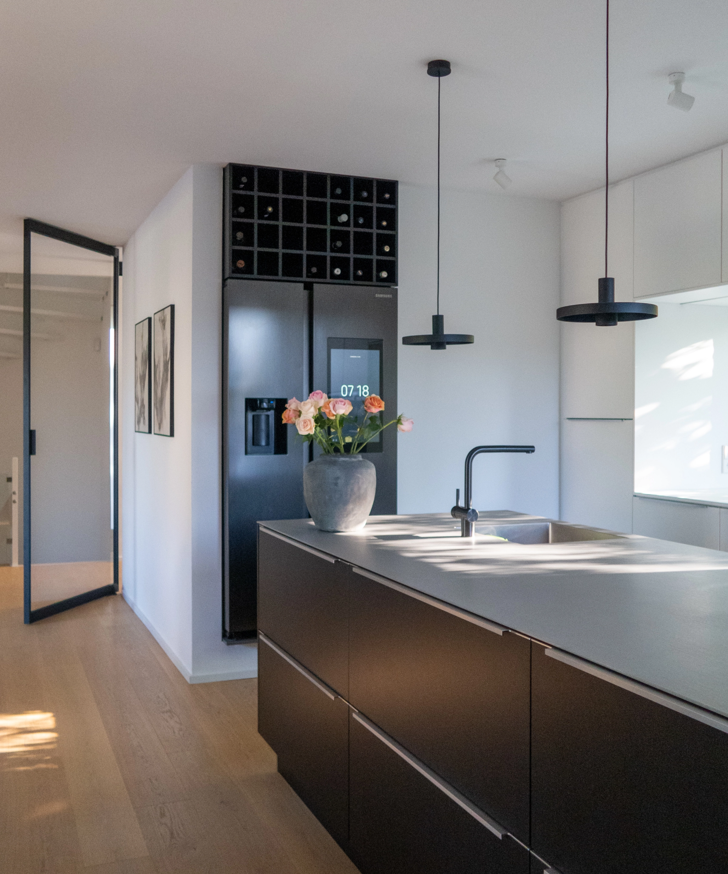 Cocina moderna con isla oscura y lámparas colgantes, refrigerador al fondo. Nolte Cocina.