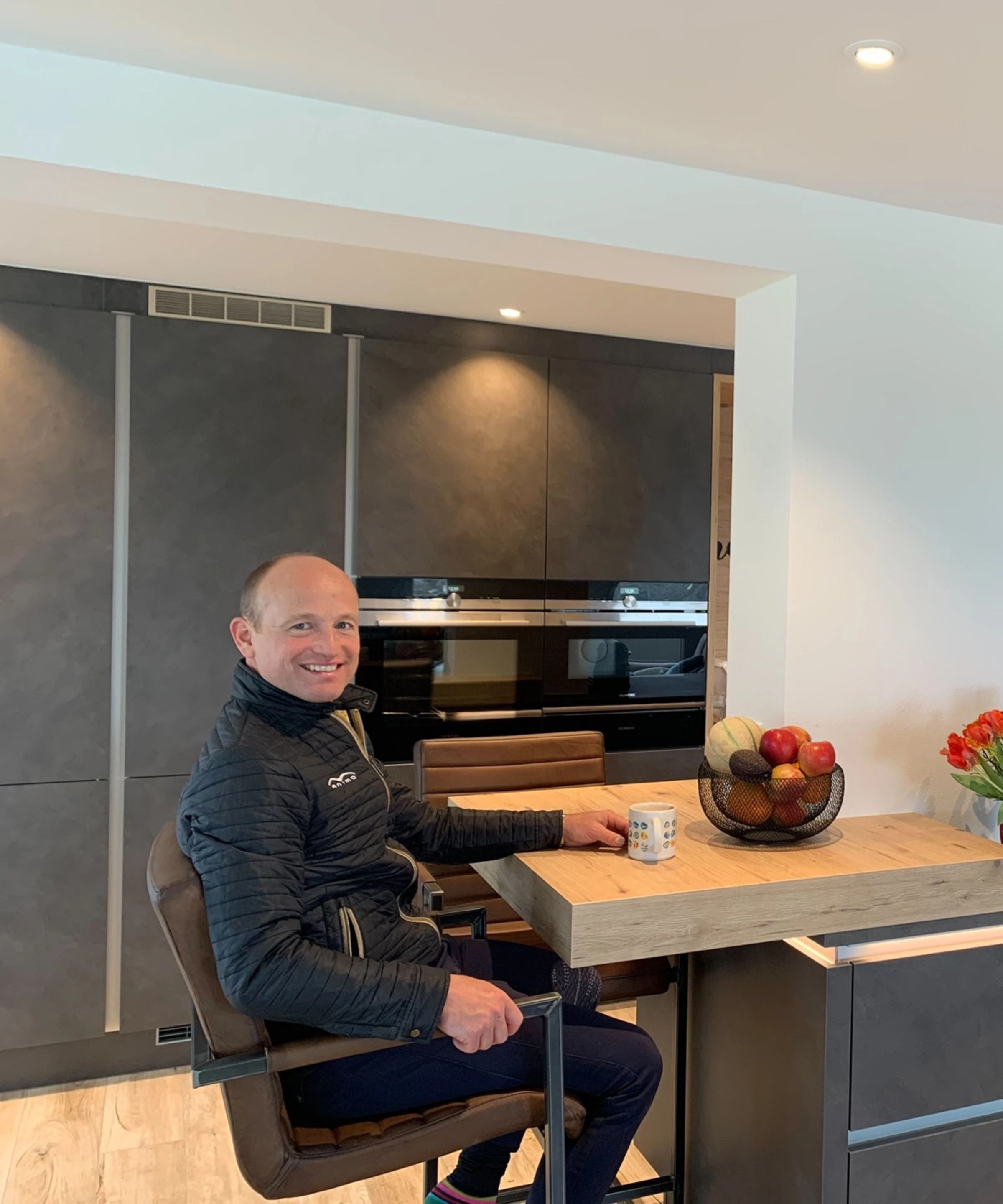 Cucina moderna con uomo al bancone, ciotola di frutta e fiori, design di Nolte Kitchen.