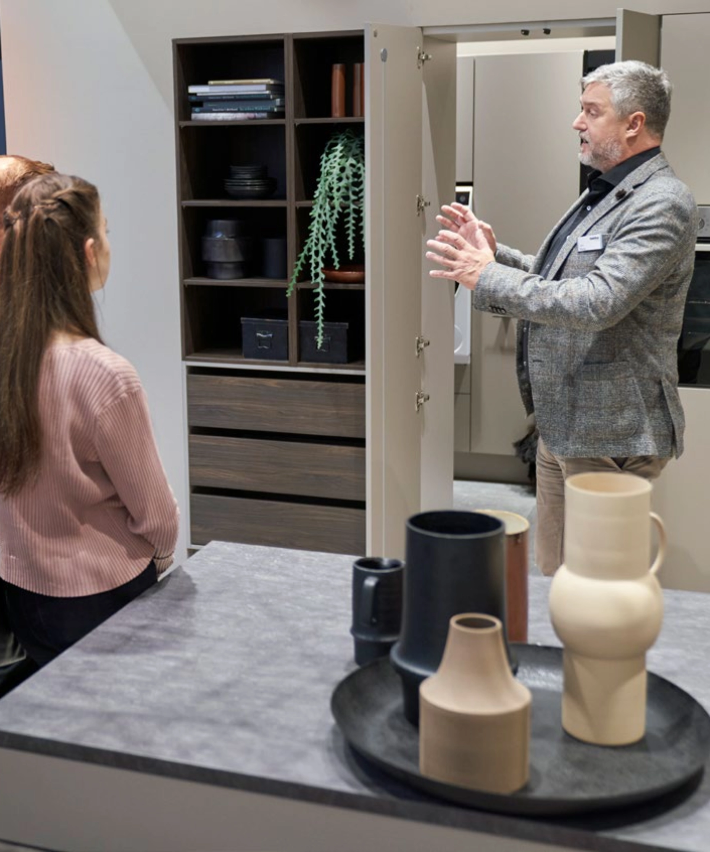 A man explains a modern kitchen with shelves, vases, and plants to two people. Nolte Kitchens.
